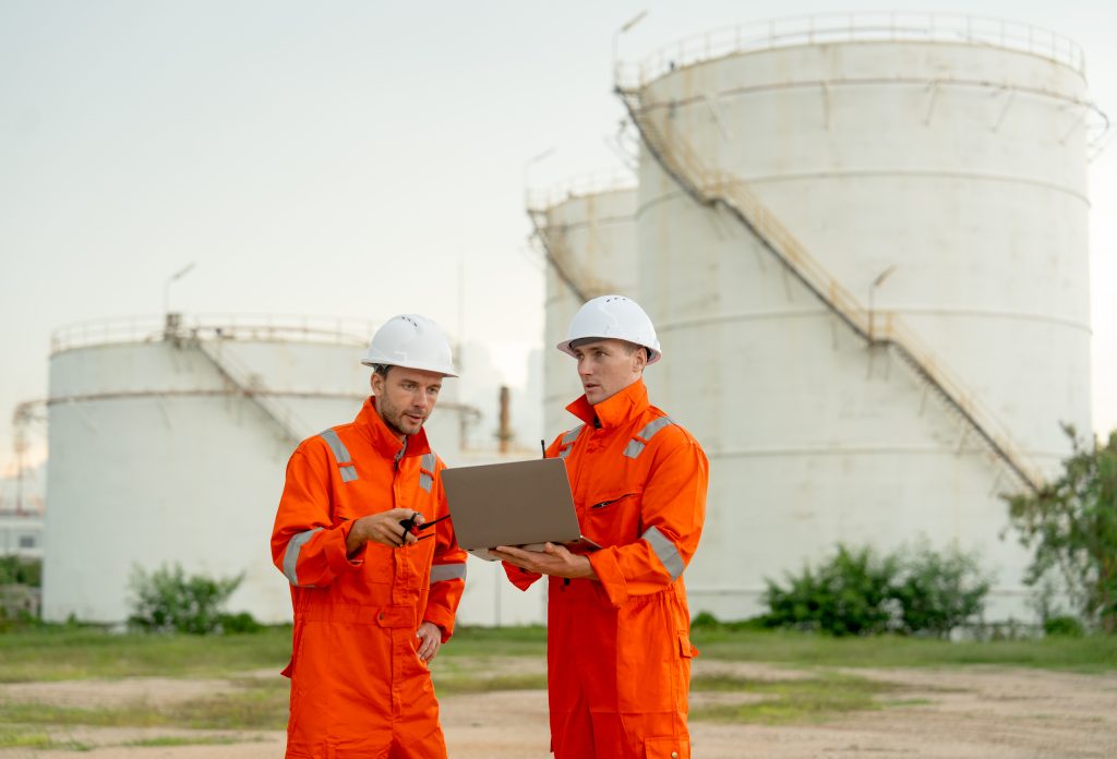 Dos ingenieros o trabajadores de una fábrica de gas conversan frente a un tanque de gas o petroquímico en su lugar de trabajo, utilizando un ordenador portátil - Antioqueña de Transportes
